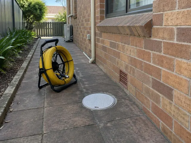 Capped drain inspection opening beside a coiled camera reel on a paved side path by a brick house in Riverwood