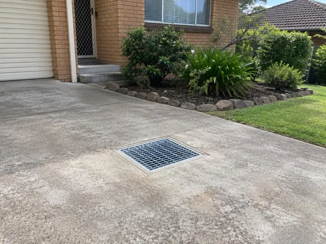 Concrete driveway with a seated stormwater grate beside a brick house and garden bed in Peakhurst