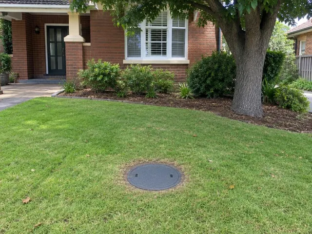 Round capped sewer access cover set in a front lawn beside a brick house in Mortdale